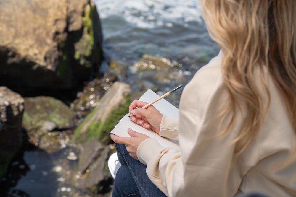 Journaling tips - Woman sitting by the water and writing in a journal.